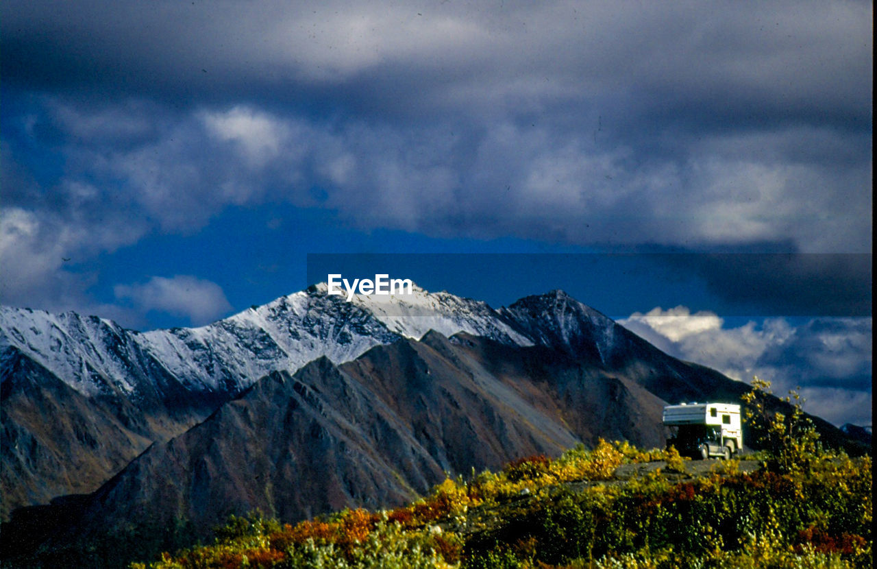 Travel trailer by snowcapped mountains against cloudy sky