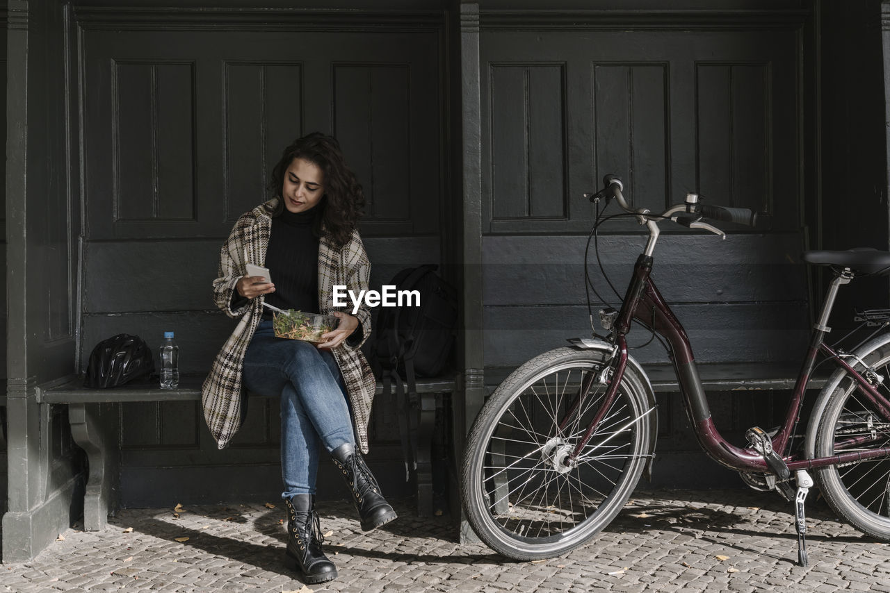 Woman with bicycle having lunch and using smartphone on station platform, berlin, germany