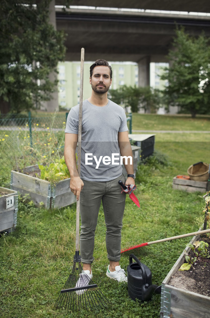 Full length portrait of mid adult man with rake and trowel in urban garden