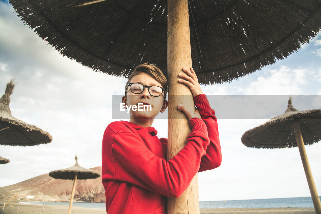 Boy standing under thatched roof at beach against sky