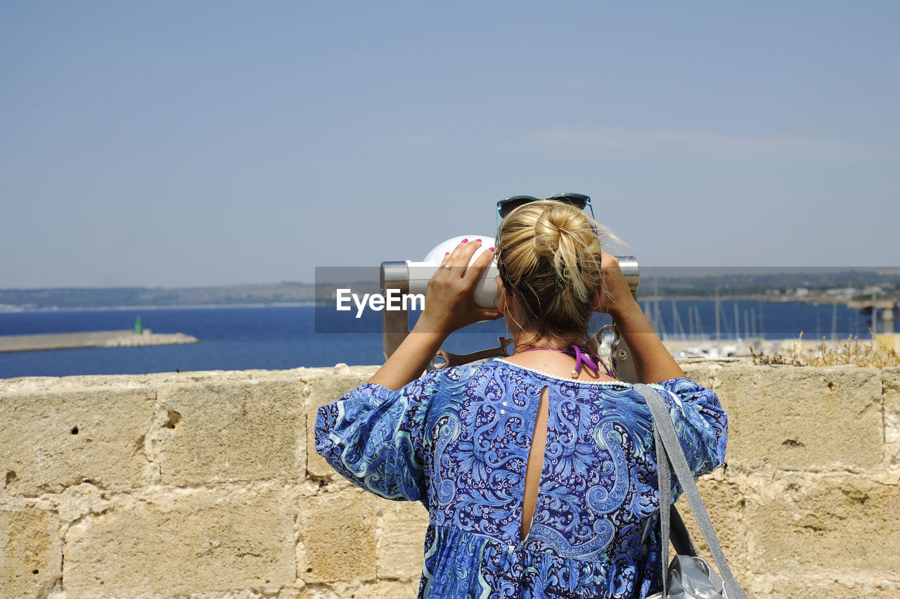 Rear view of woman looking through coin-operated binoculars against sky