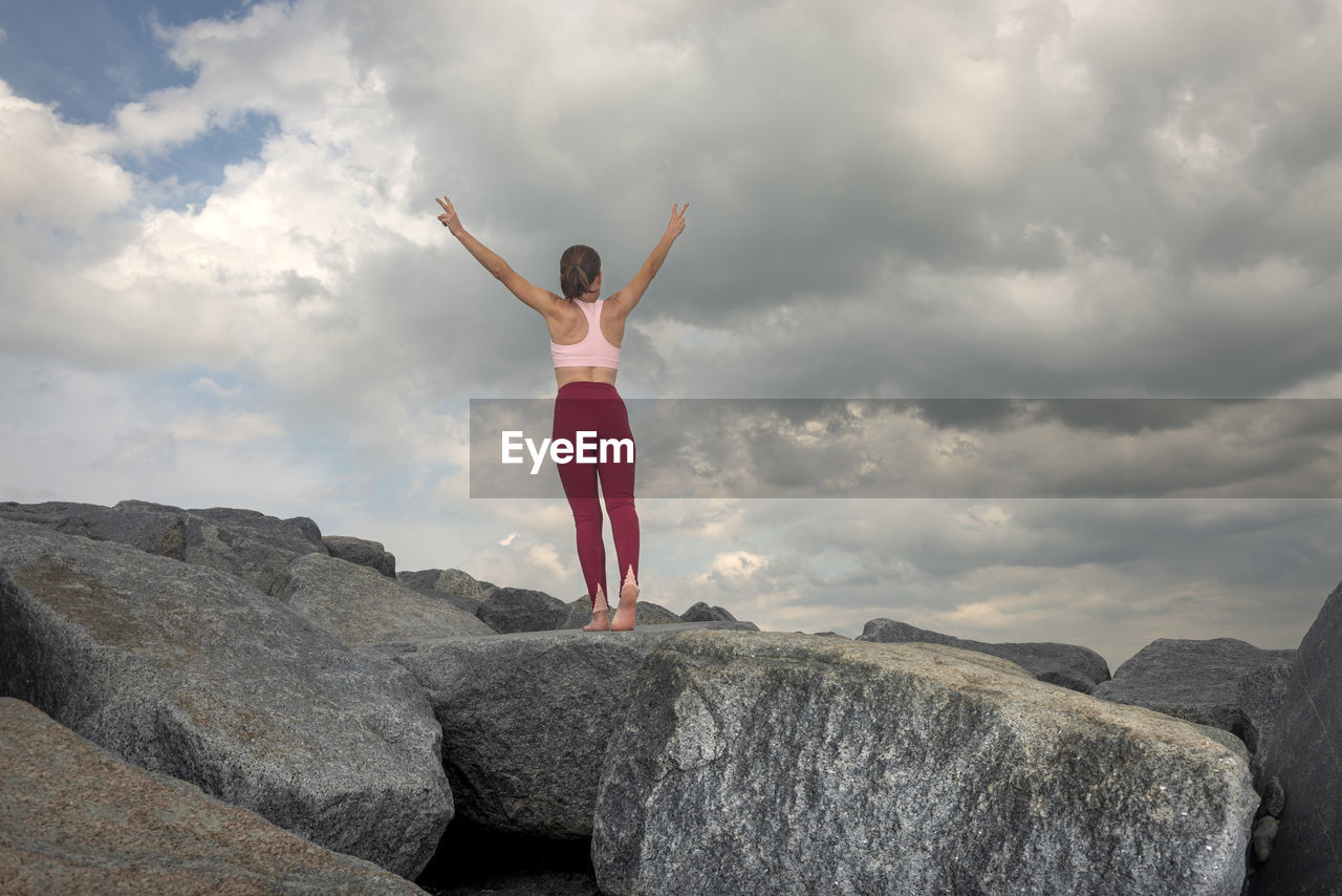 Sporty woman stands on top of rocks after a climb, arms raised in celebration.