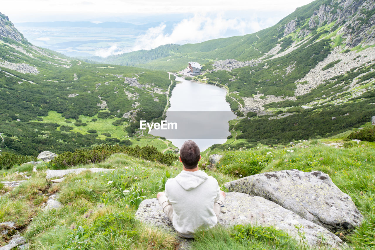 Rear view of man sitting on rock against river and mountains