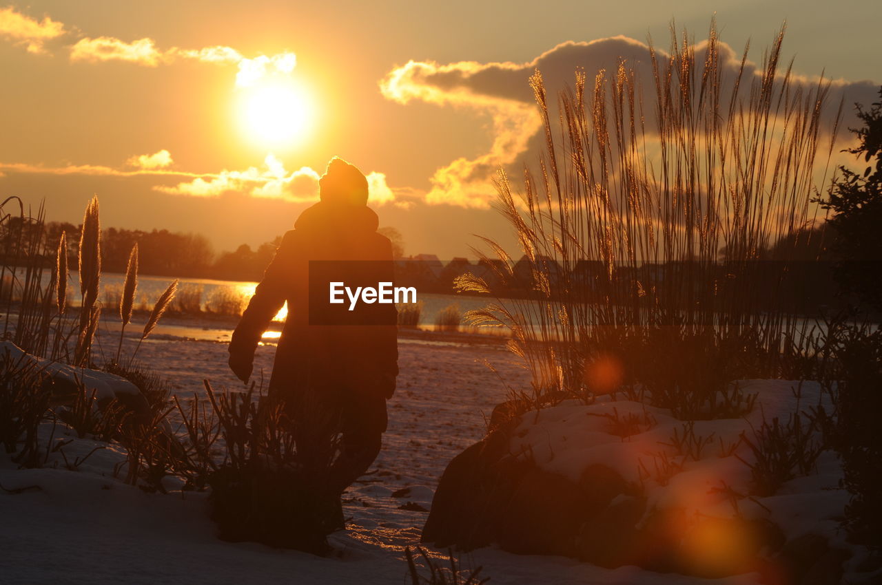 REAR VIEW OF SILHOUETTE MAN STANDING AT SHORE DURING SUNSET