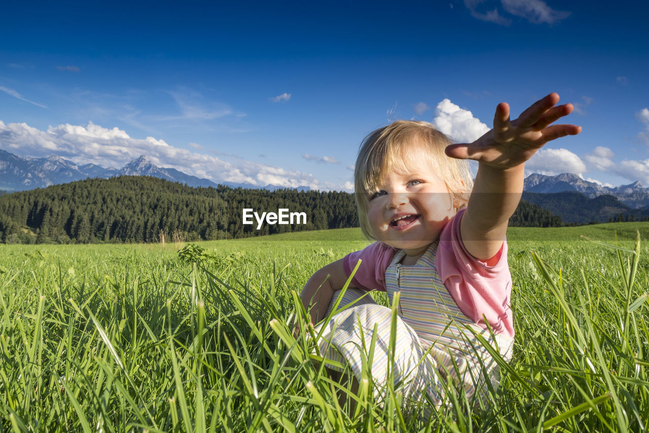 Smiling cute girl gesturing while sitting on grass against sky