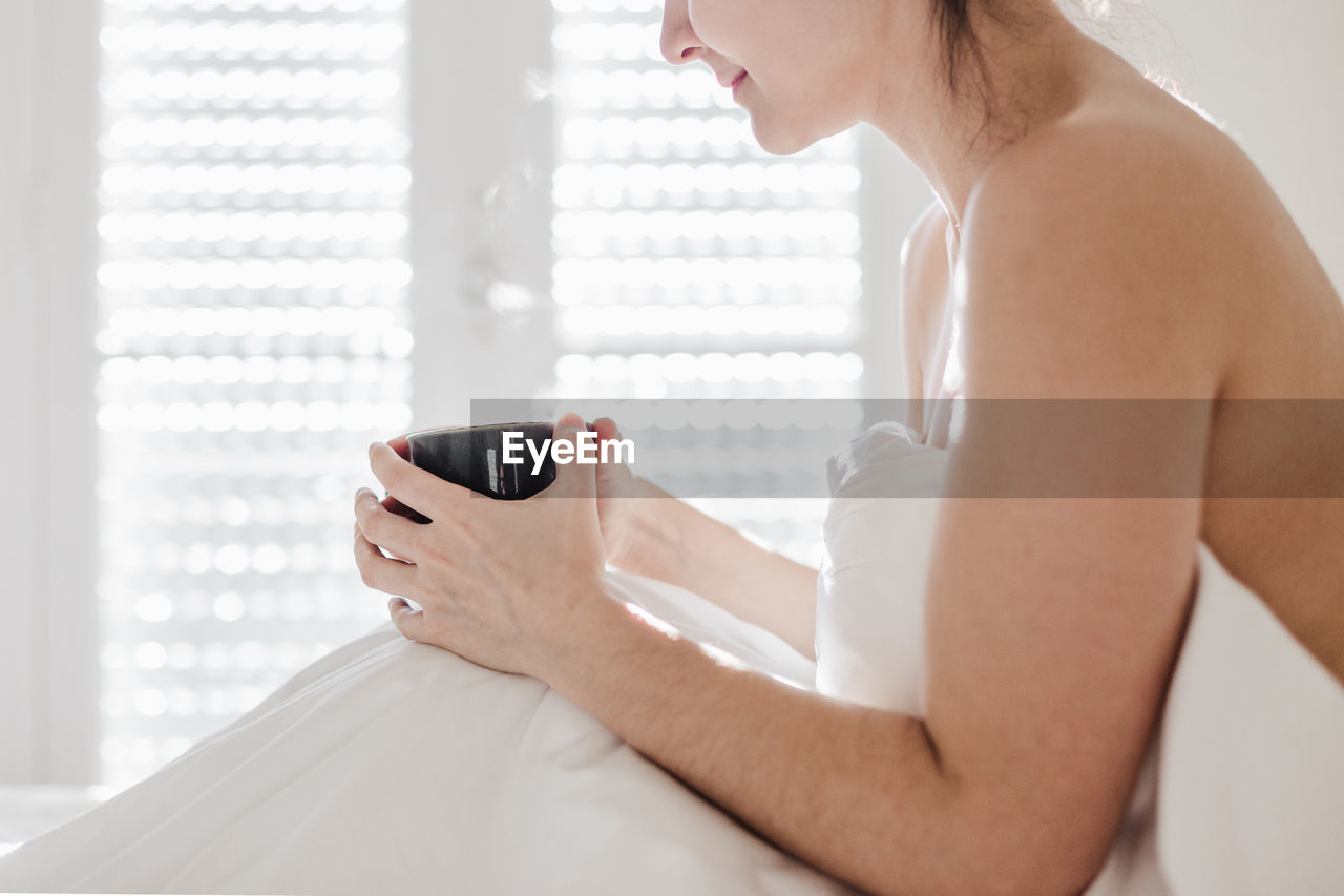 SIDE VIEW OF WOMAN HOLDING COFFEE CUP AT HOME