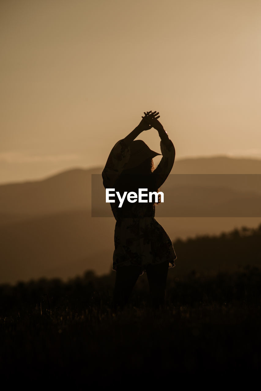 Woman standing with arms raised on land during sunset