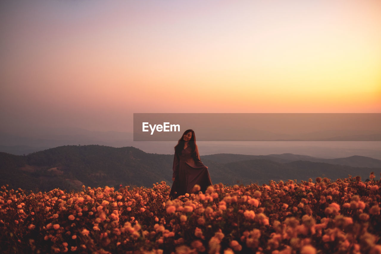 Beautiful woman standing amidst field against sky during sunset