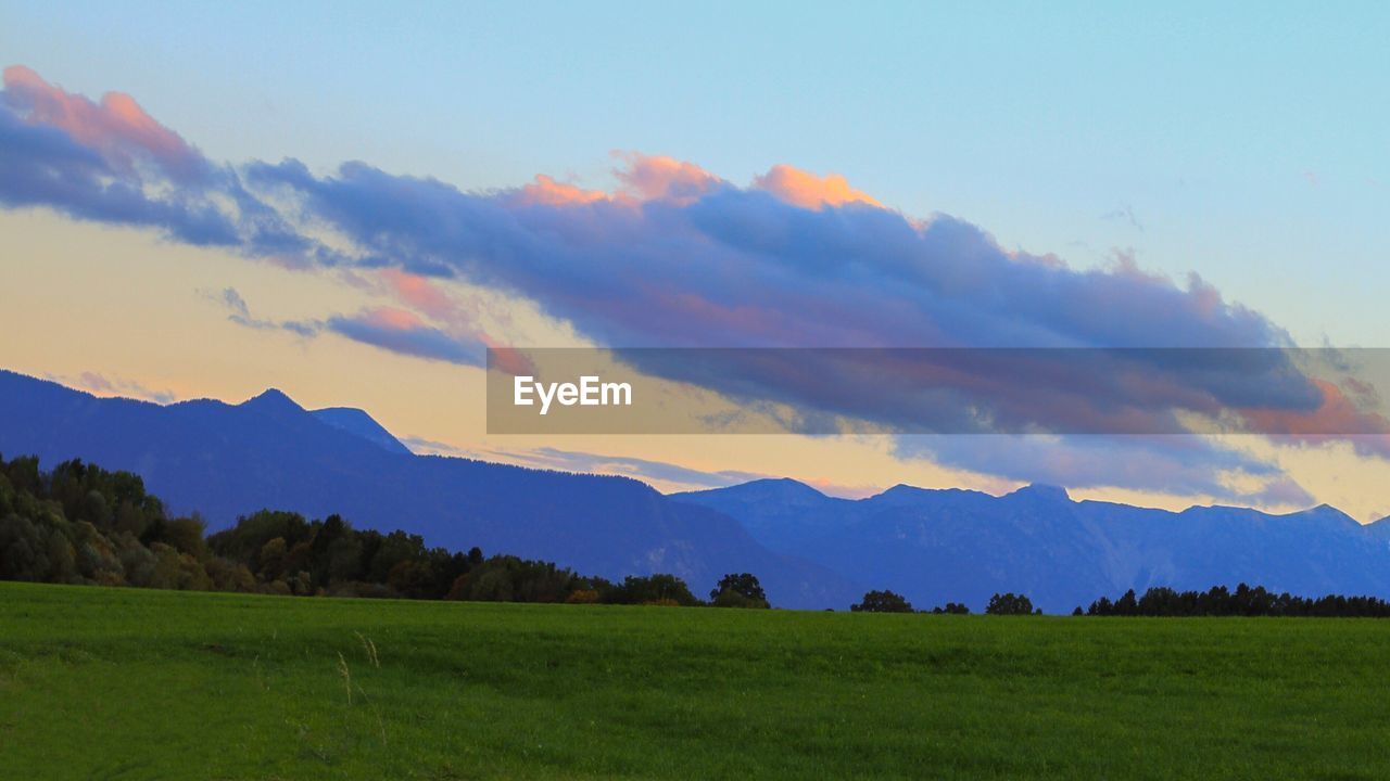 SCENIC VIEW OF GRASSY LANDSCAPE AGAINST MOUNTAINS AT SUNSET