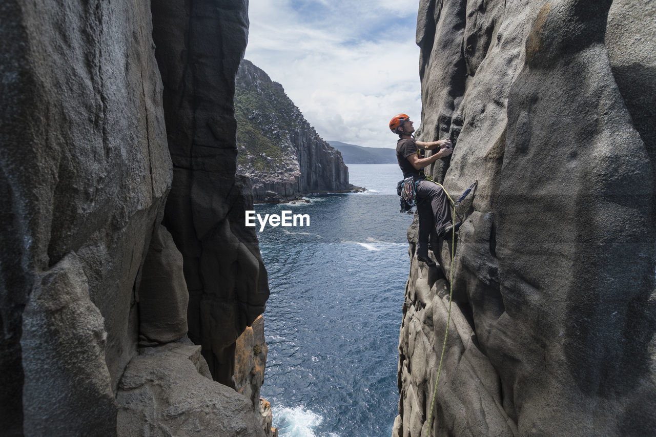 Daring man rockclimbs the exposed edge of a dolerite rock ridge, with the ocean seen in the background in cape raoul, tasmania, australia.