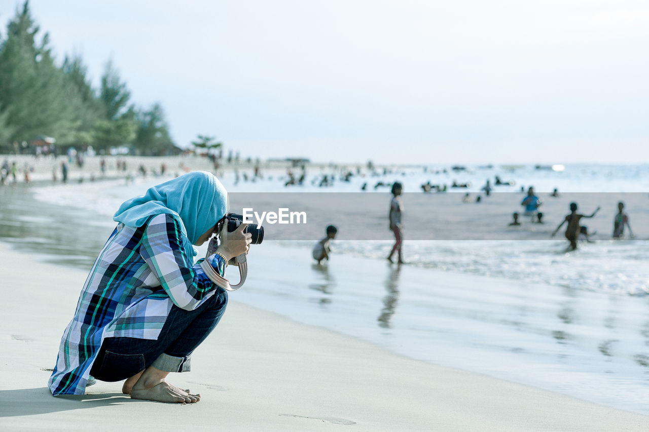 Side view of woman photographing while crouching against people at beach