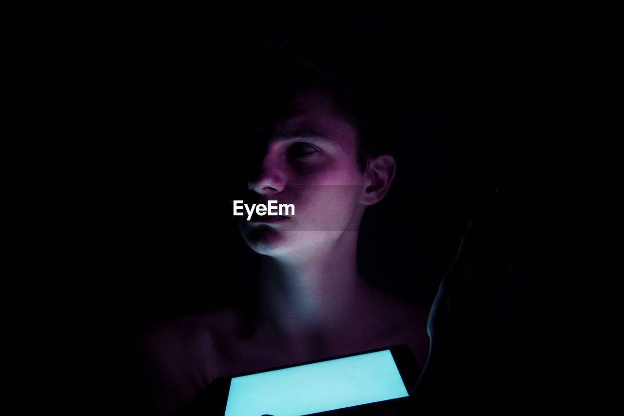 Close-up of teenage boy with illuminated mobile phone looking away in darkroom