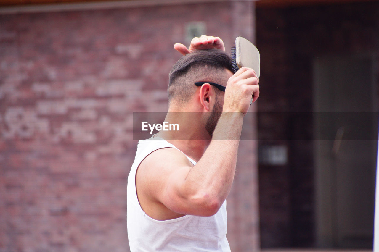 Side view of man brushing hair while standing against wall