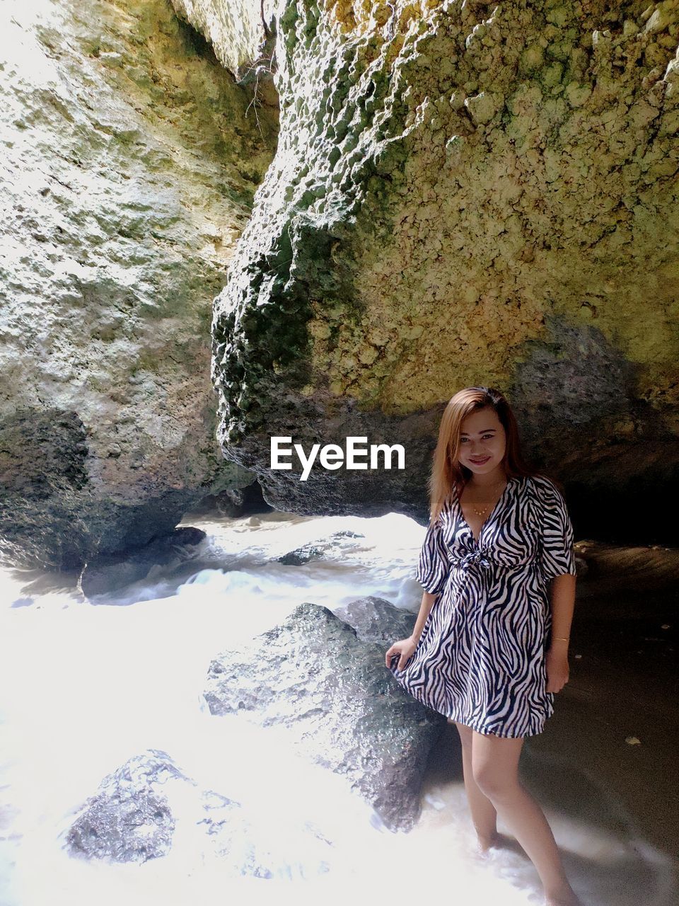 FULL LENGTH OF YOUNG WOMAN STANDING ON ROCK AGAINST WATERFALL