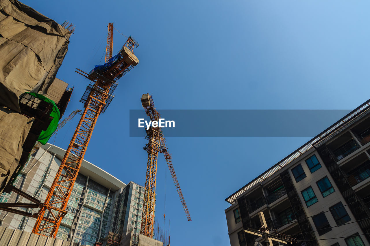 LOW ANGLE VIEW OF CRANE BY BUILDINGS AGAINST CLEAR BLUE SKY