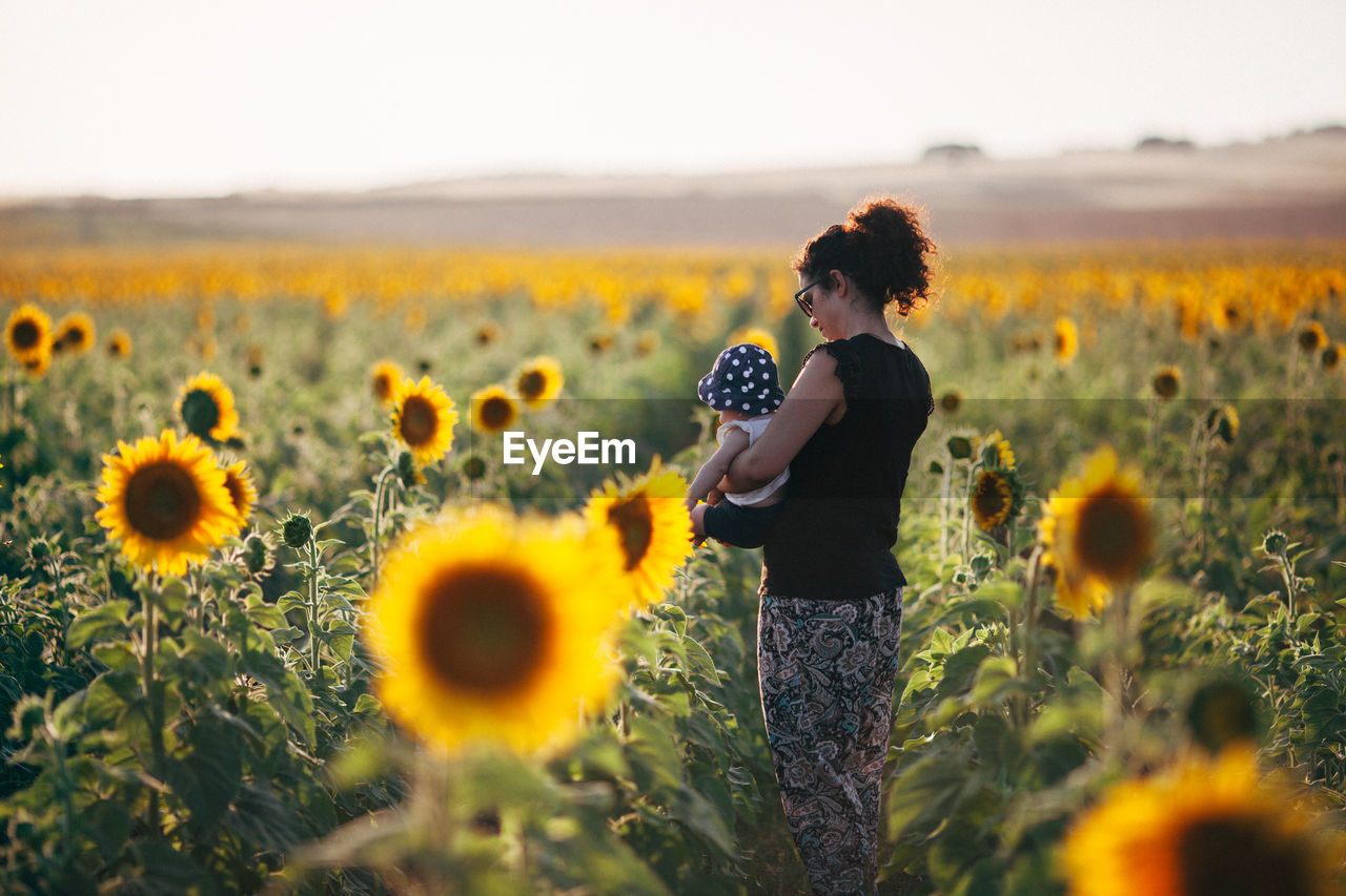 Side view of mother carrying daughter while standing amidst sunflowers in field against sky