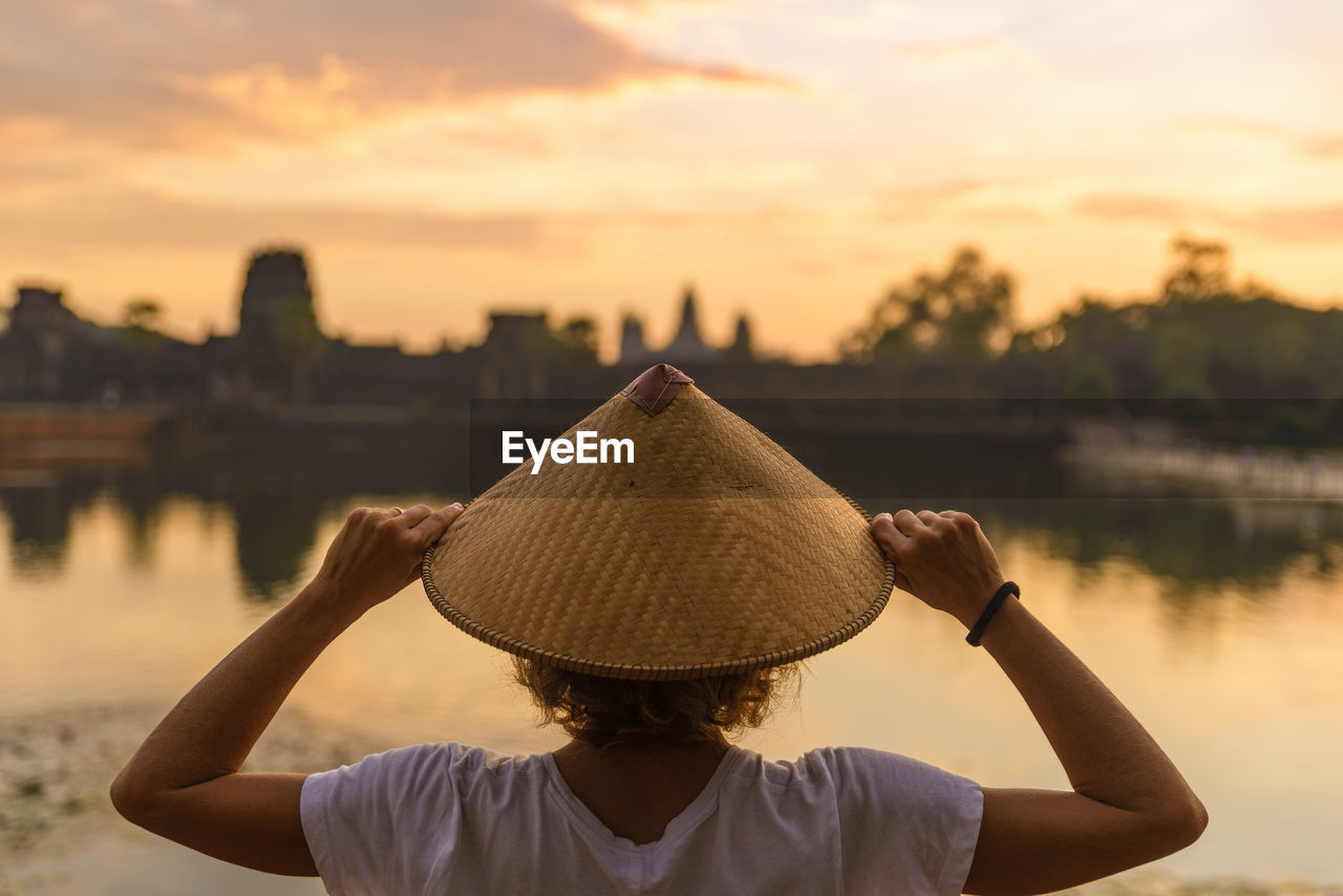 Rear view of woman wearing hat against lake during sunset