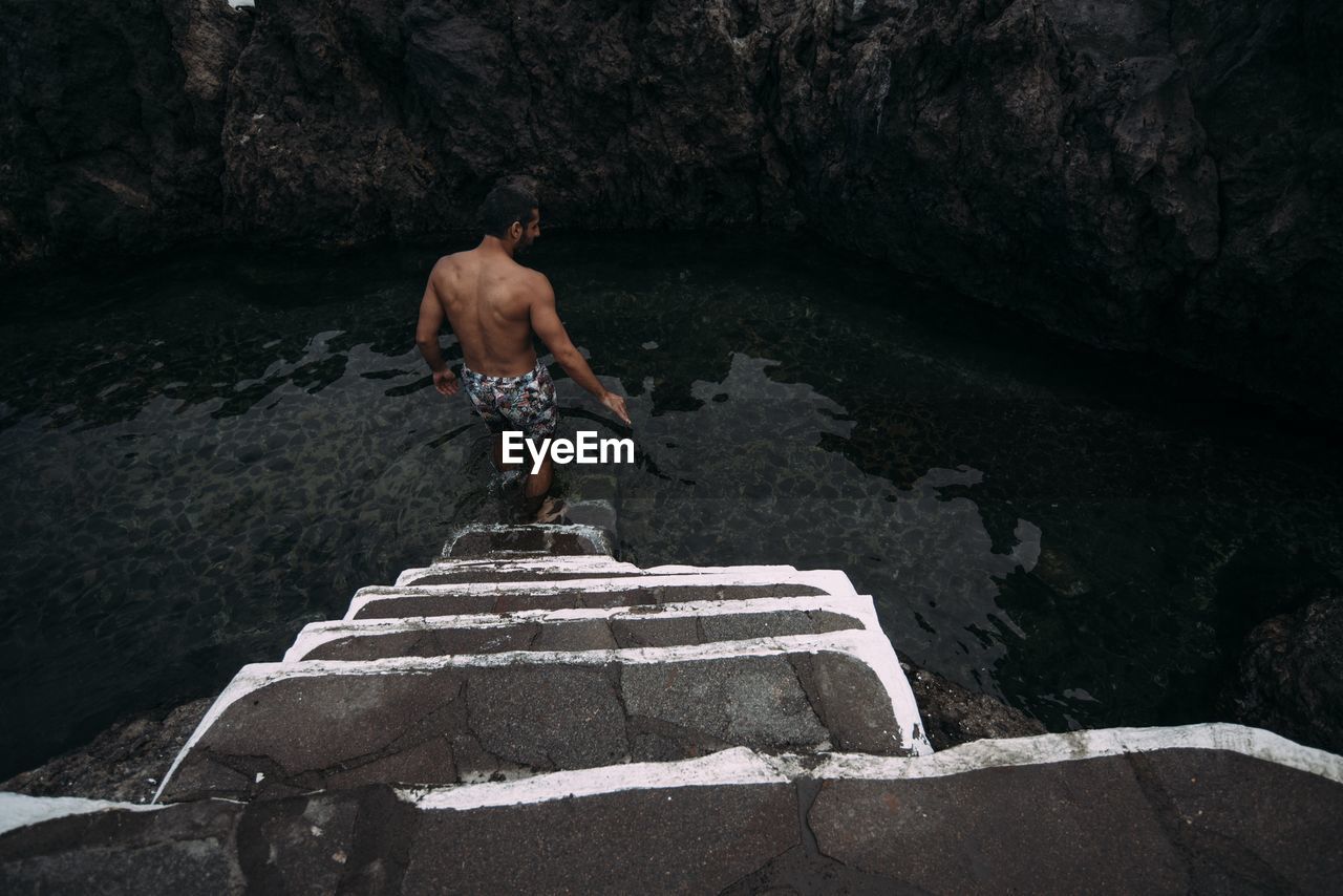 REAR VIEW OF SHIRTLESS MAN STANDING ON ROCK AGAINST WATERFALL