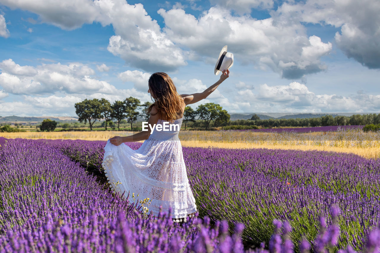 Woman seen from behind plays with her long white dress in the middle of a blooming lavender field.