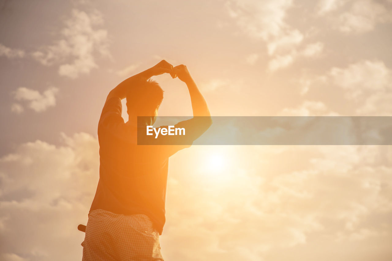 Rear view of man with arms raised standing against sky during sunset