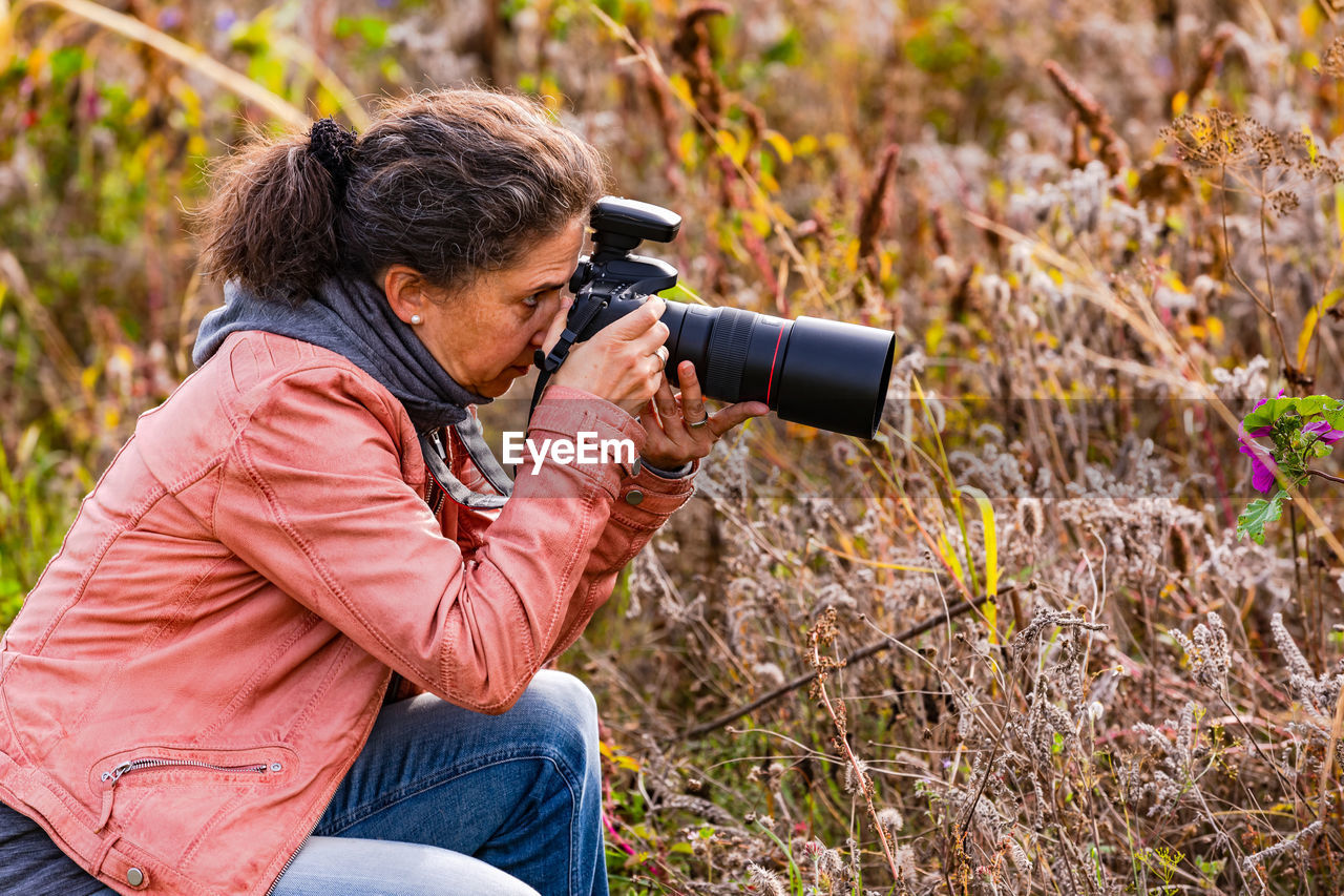 Side view of an attractive woman with professional dslr camera in meadow photographing wild flowers