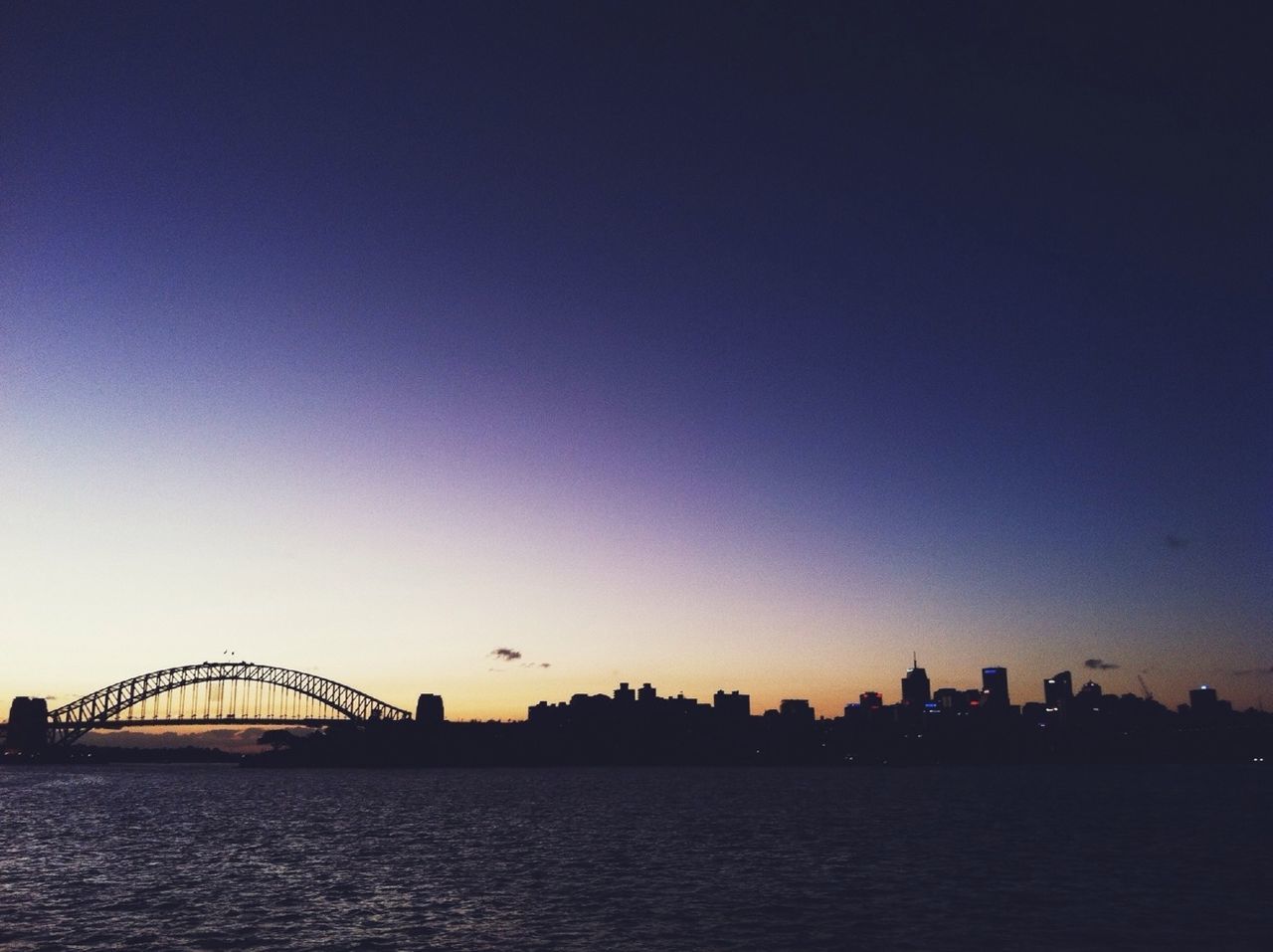 Silhouette bridge over river by cityscape against sky at dusk