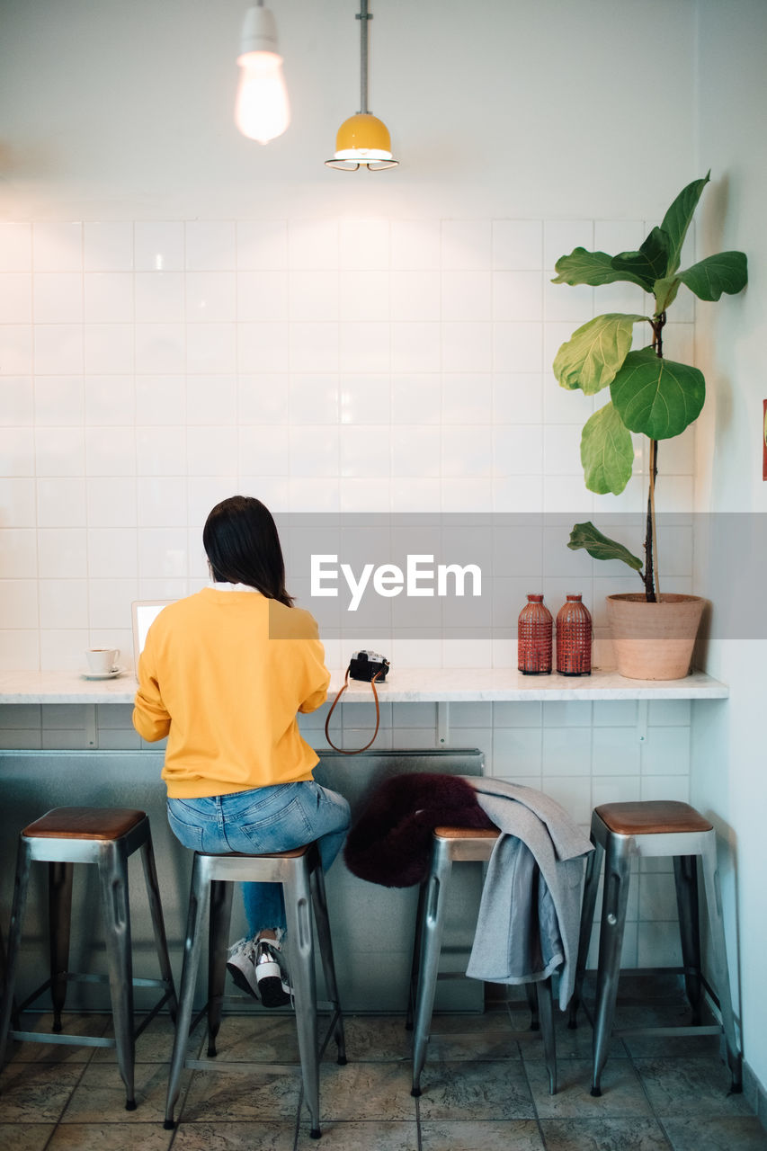 Full length rear view of female blogger sitting on stool at cafe