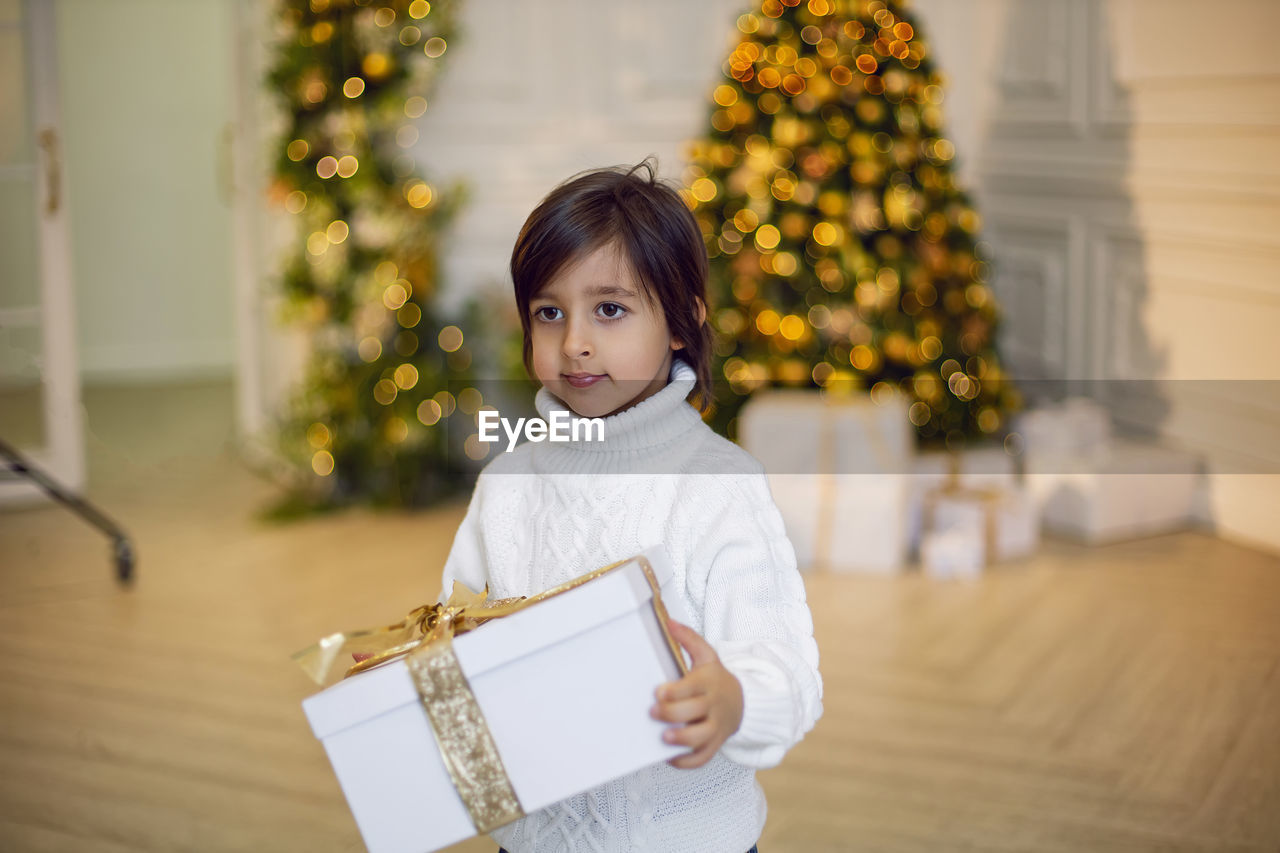 Portrait of a boy a child in a white sweater holding a gift box at the christmas tree