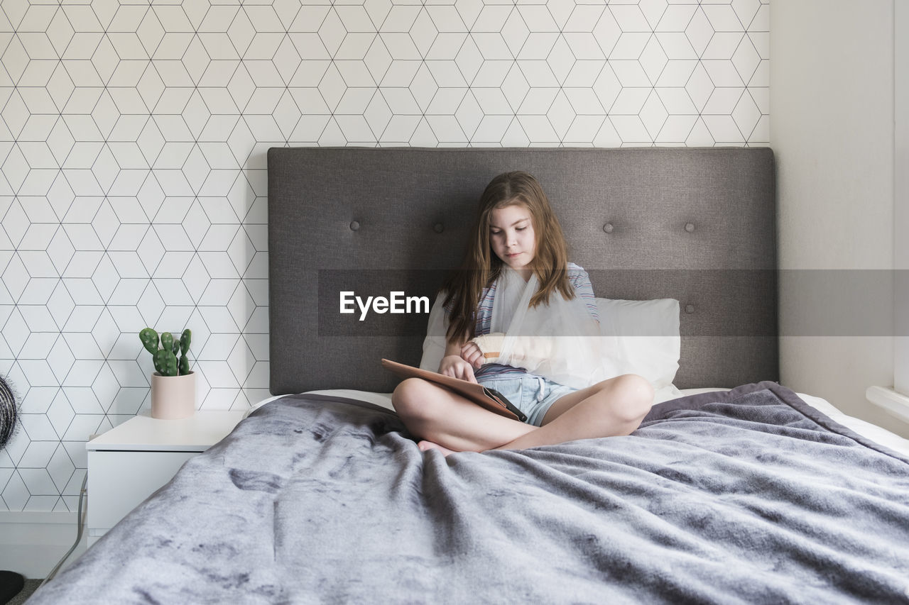 Young girl sitting on bed at home with a broken arm in a sling