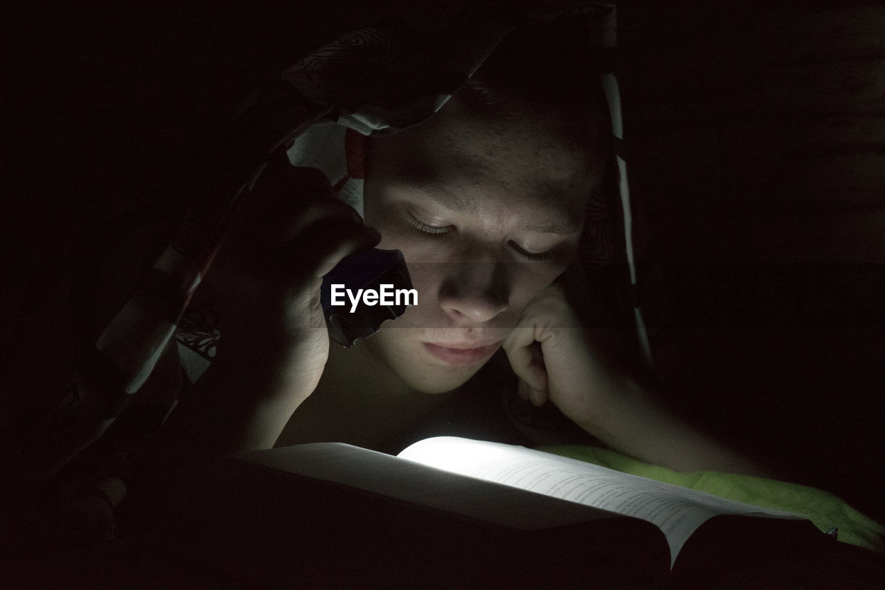 A boy reading a book in the dark by lighting the pages with a flashlight