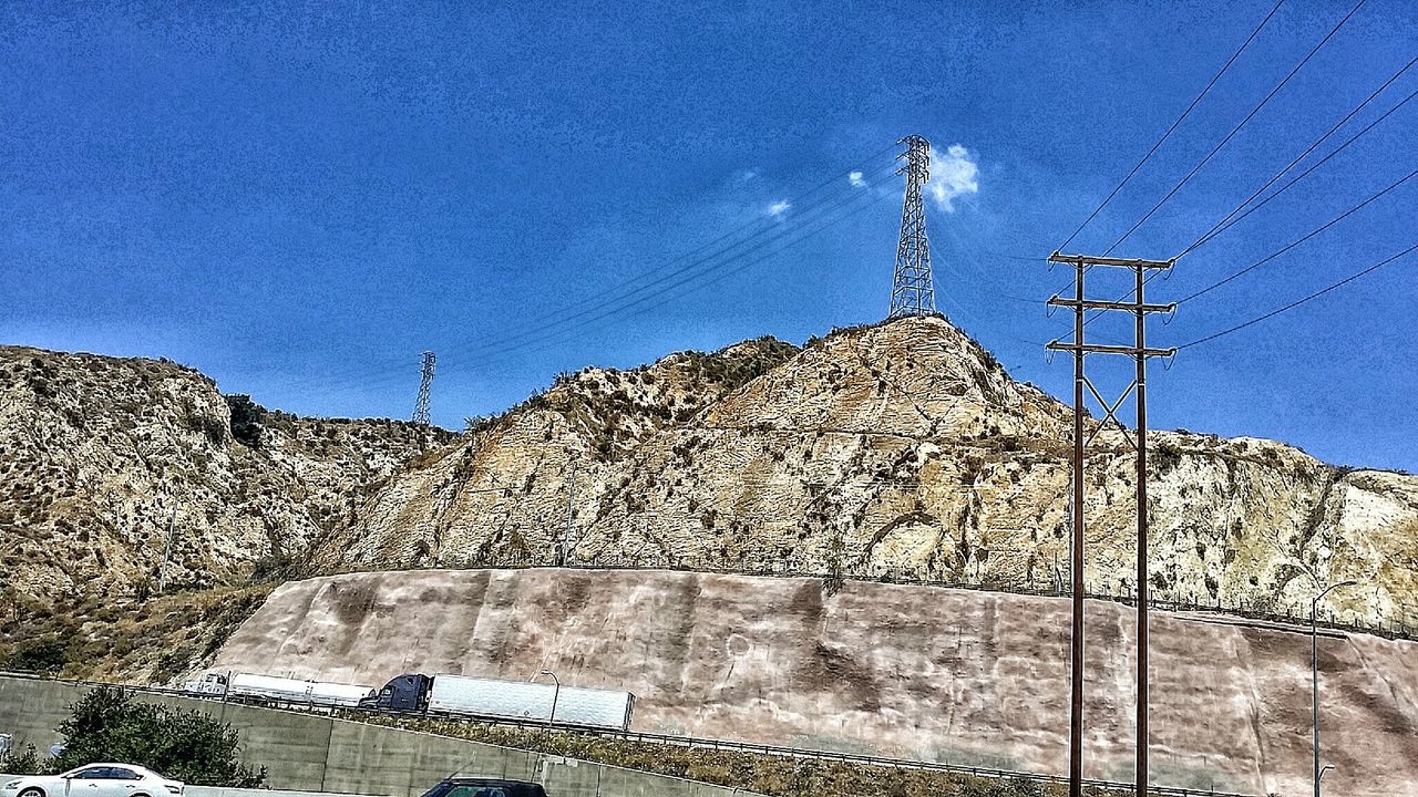 Low angle view of electricity pylons on mountains against blue sky