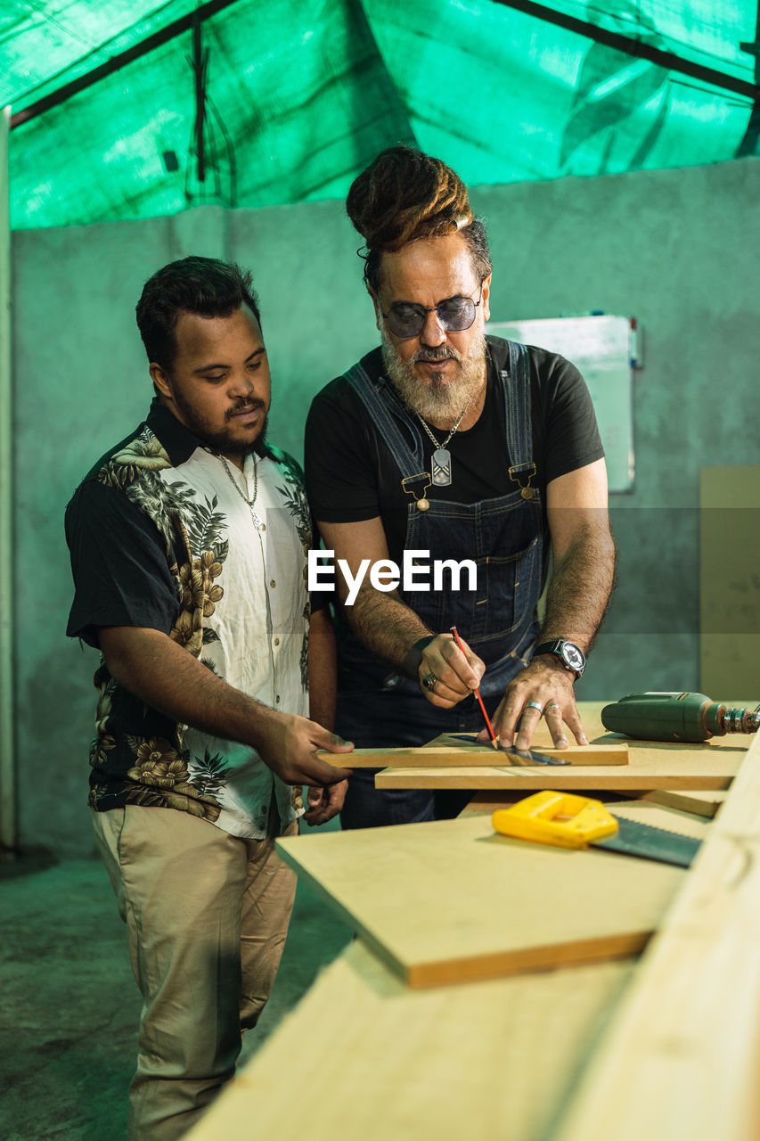 Serious multiracial male colleagues with ruler measuring wooden plank while standing at table with saw in professional joinery with special equipment