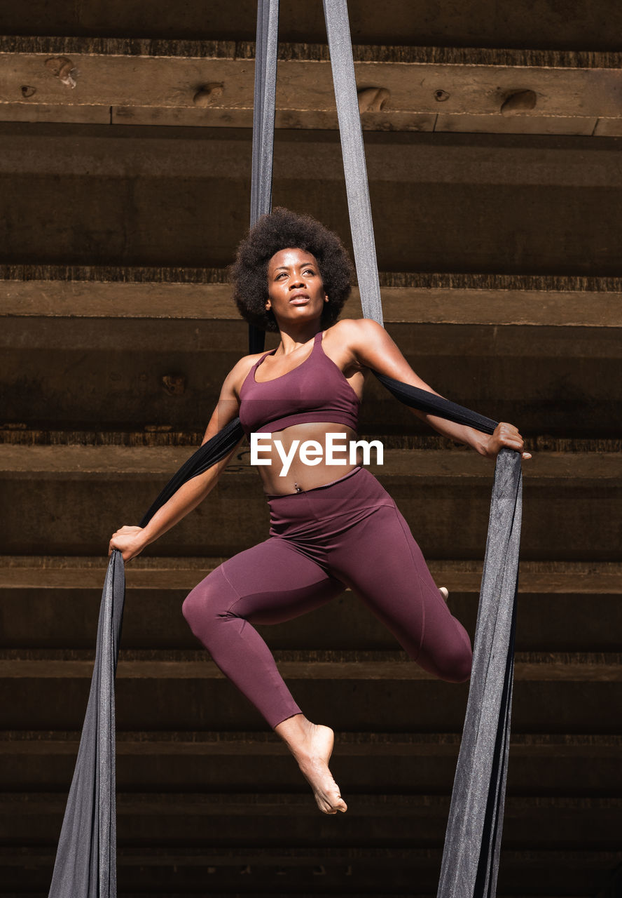 Slender african american female balancing on hanging hammock while doing aerial yoga above ground under bridge in city