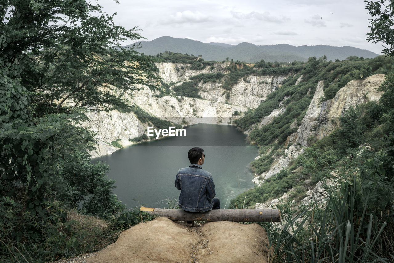 REAR VIEW OF MAN SITTING ON MOUNTAIN AGAINST TREES