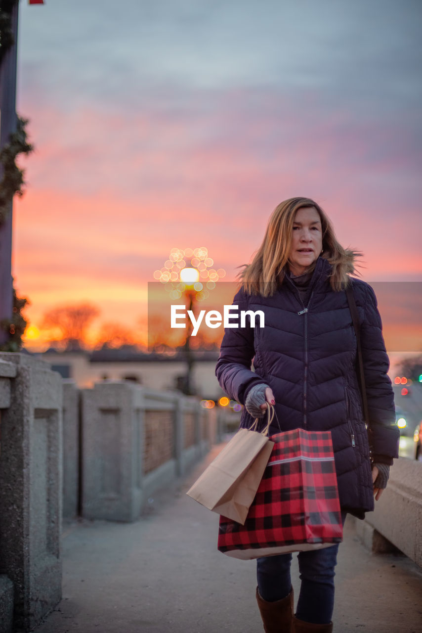 Woman looking away while walking on footpath against sky during sunset