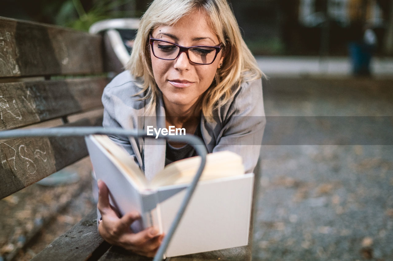 Young woman reading book while lying on bench in park