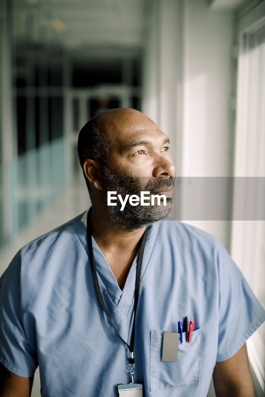 Contemplating male nurse looking through window while standing in hospital corridor