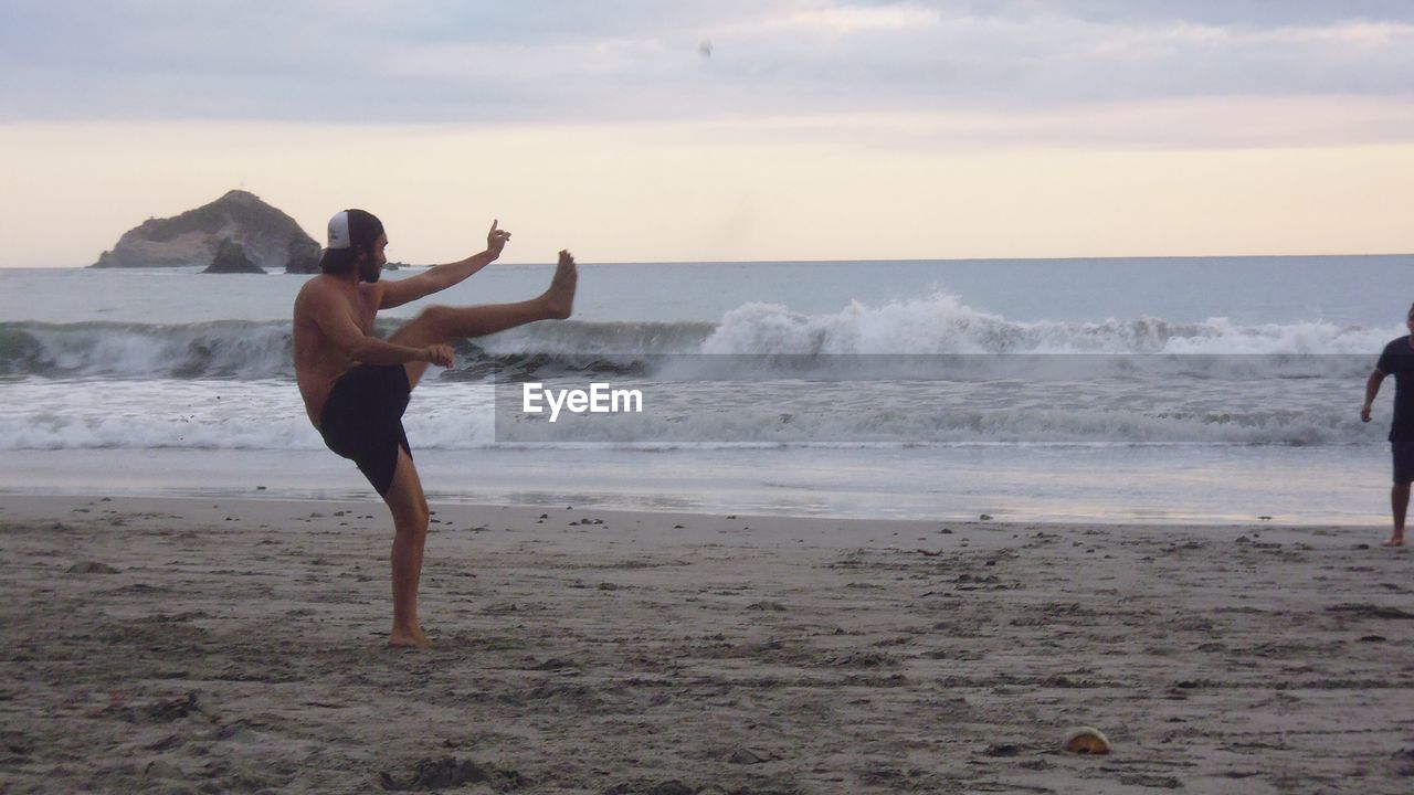 Side view of shirtless young man playing at sandy beach during sunset