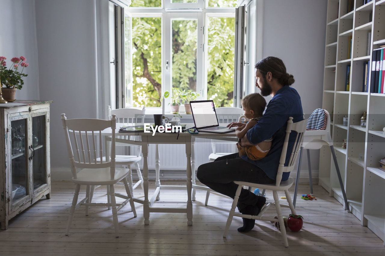 Full length of father using laptop computer while taking care of baby at home