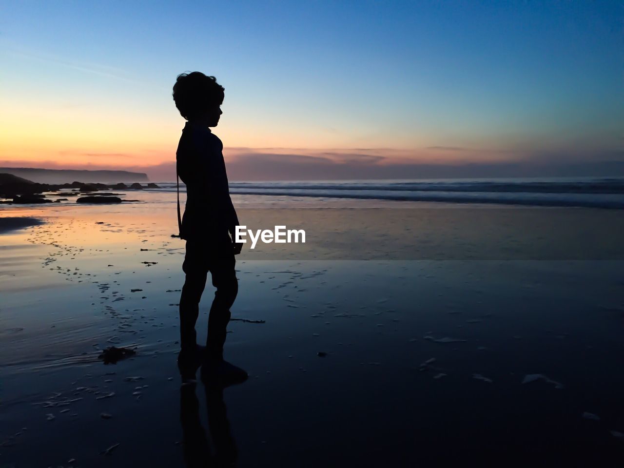 Silhouette boy standing on shore against sky during sunset