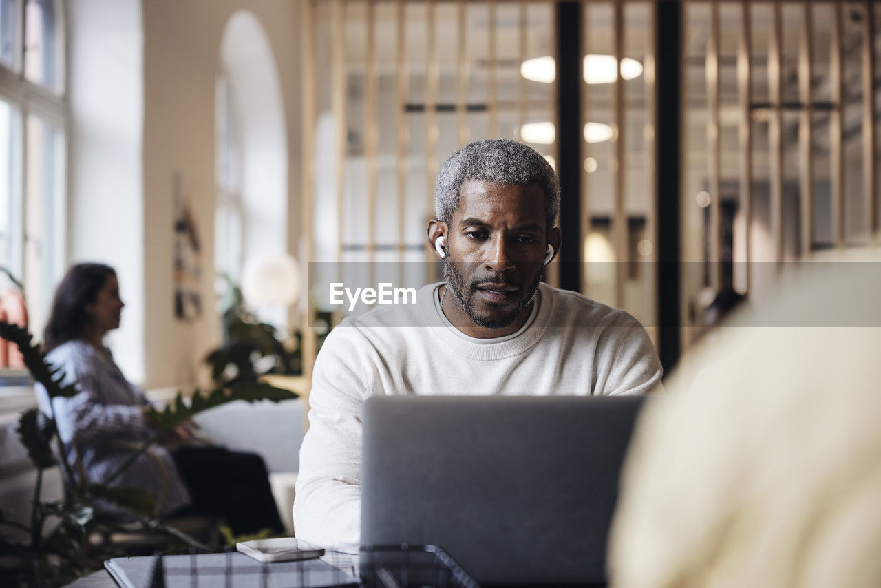 Mature male business professional working on laptop while wearing wireless in-ear headphones at coworking office