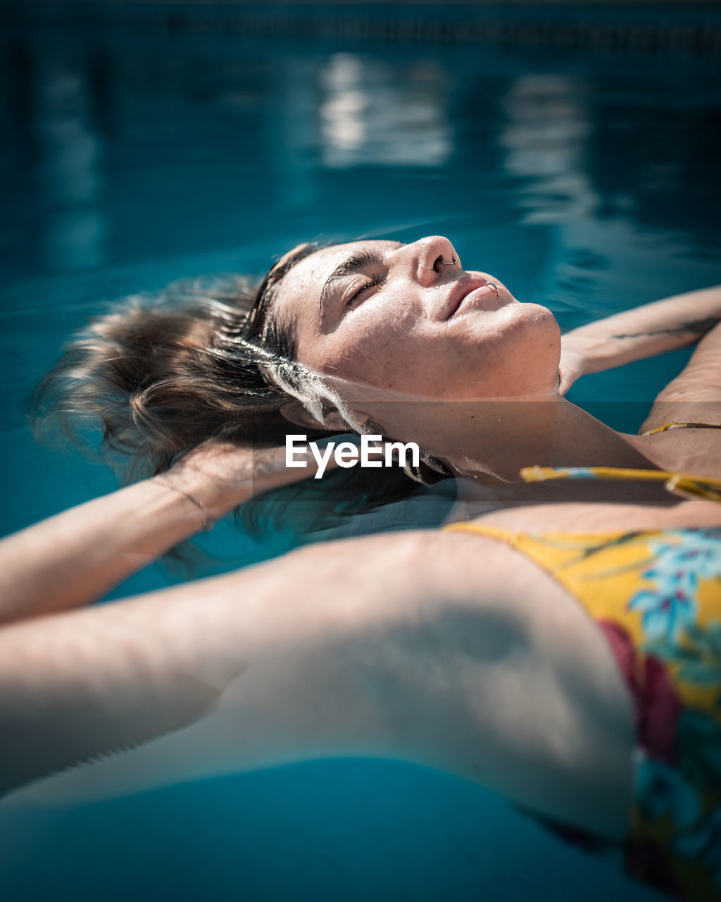 Content young female in swimwear touching head and floating with eyes closed in warm pool water on sunny day