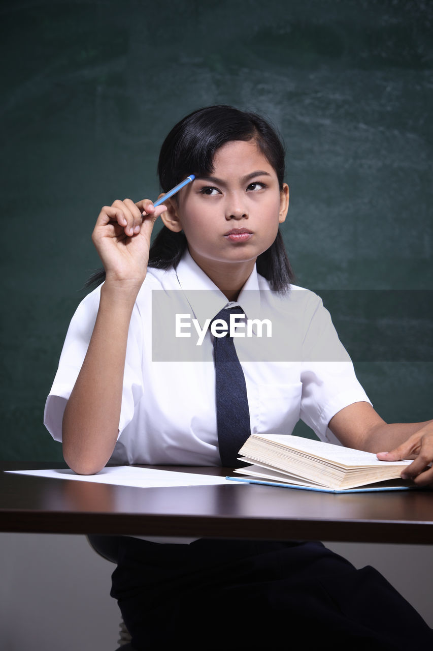 Schoolgirl thinking while studying at table against blackboard