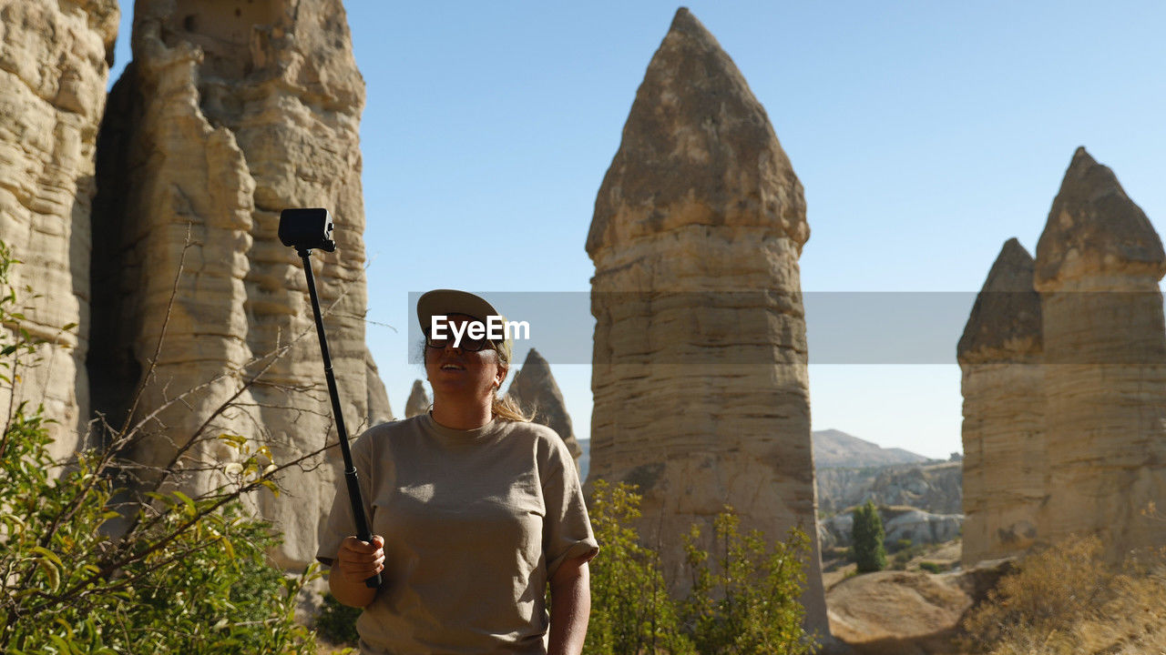 A woman films herself on an action camera against a backdrop of sandstone cliffs.  girl is blogging