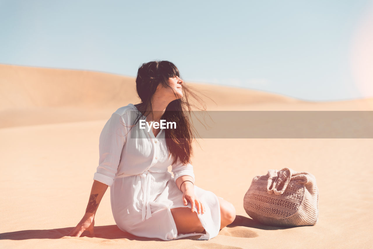 Young woman sitting on sand at beach against clear sky