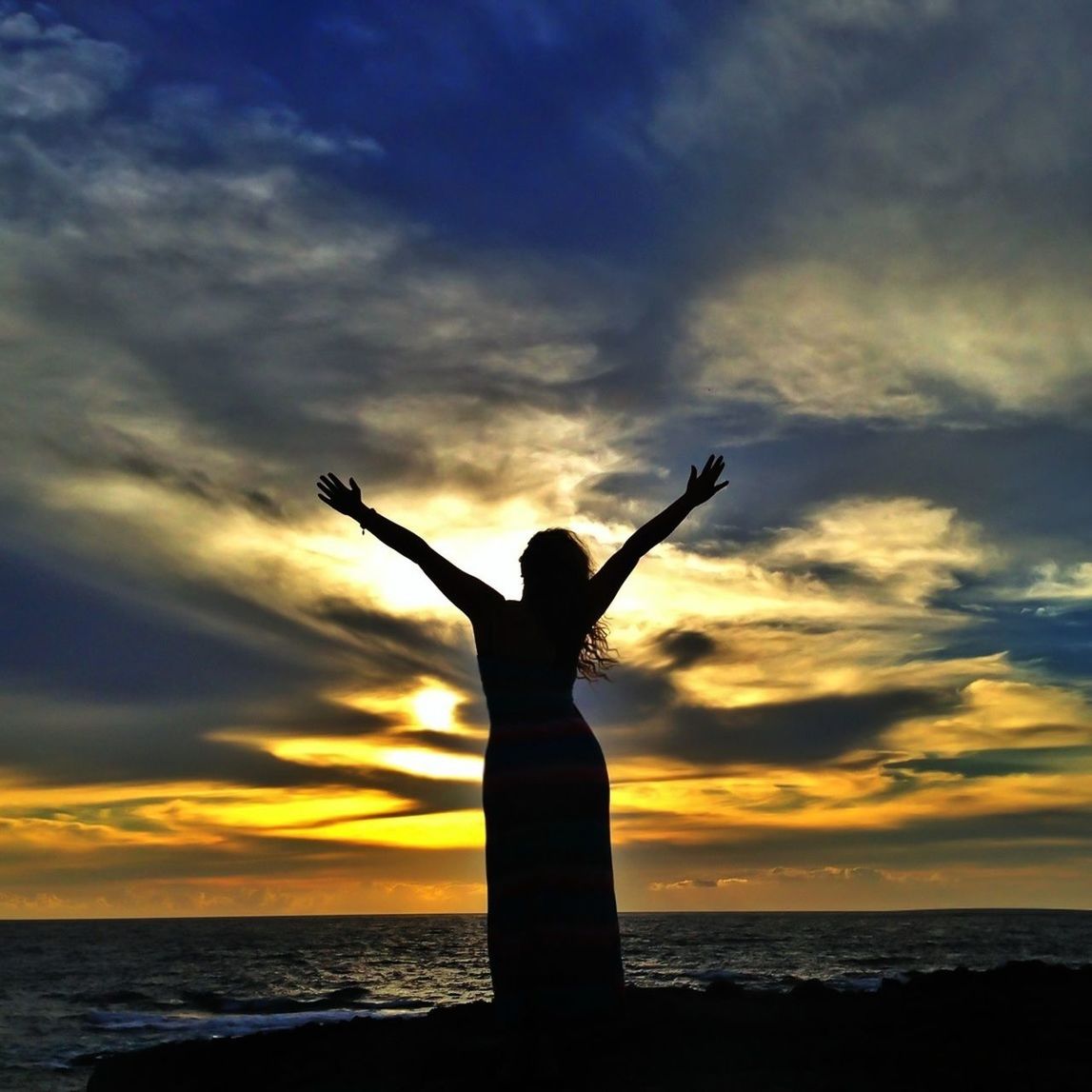 Silhouette of woman with arms outstretched standing on beach at sunset