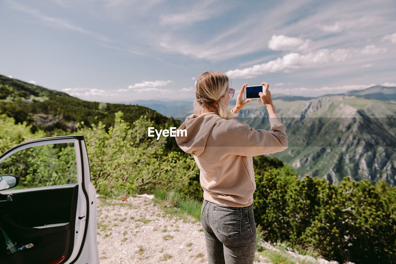 MAN PHOTOGRAPHING SMART PHONE AGAINST MOUNTAINS