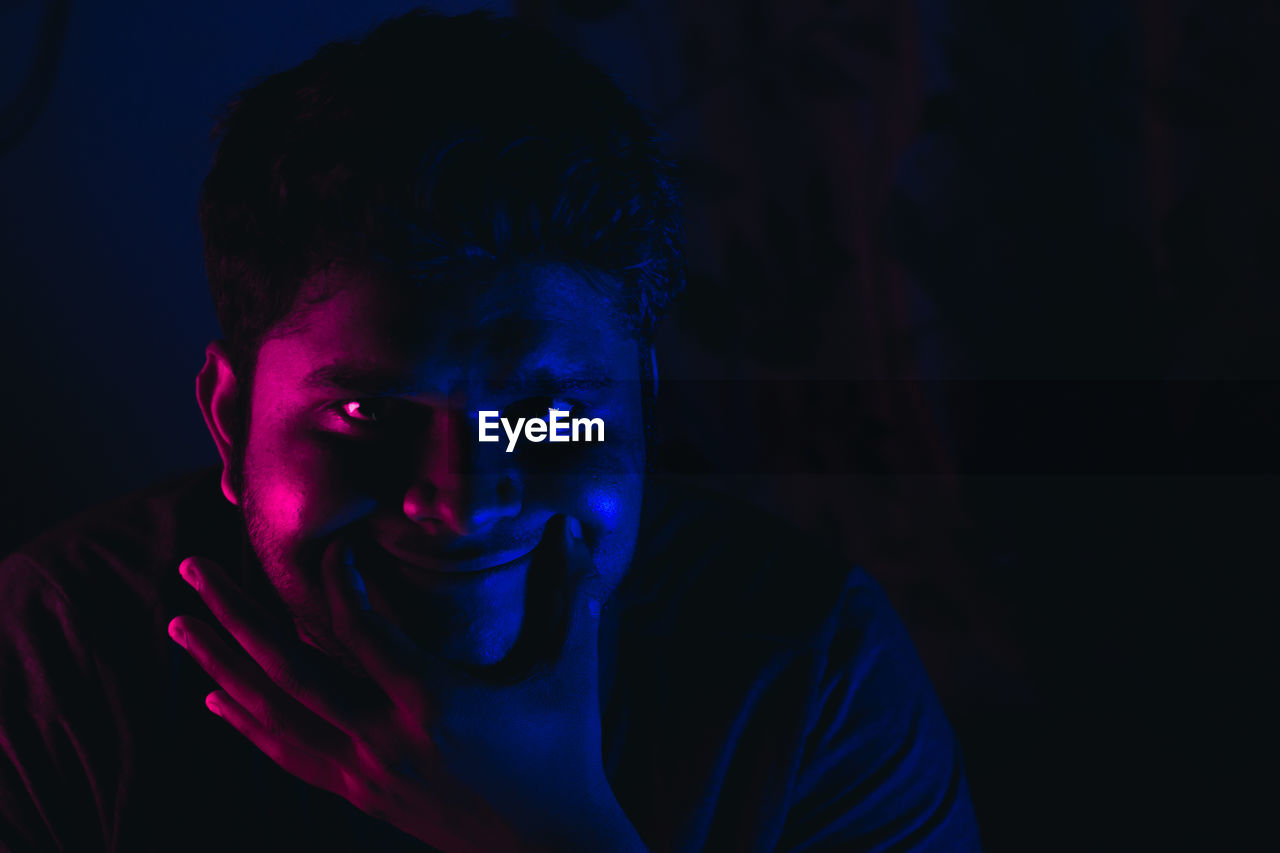 Close-up of young man smiling in multi colored lights against black background