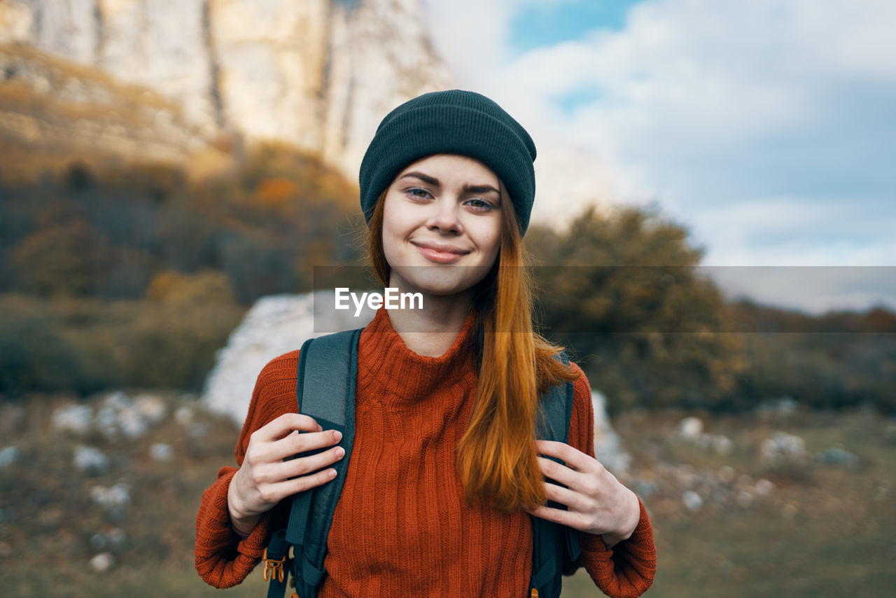 PORTRAIT OF SMILING YOUNG WOMAN WEARING HAT STANDING AGAINST TREES