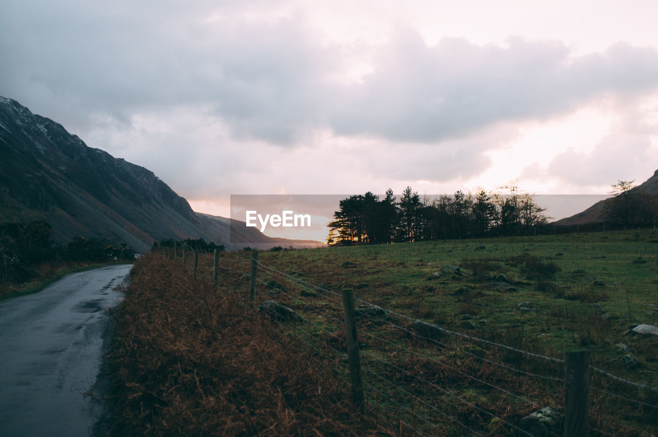 Single lane road on field by mountain against cloudy sky